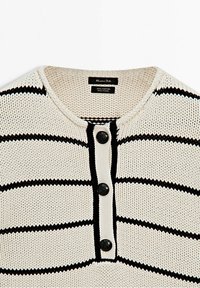 Pull en maille crème avec rayures noires horizontales, col rond et trois boutons noirs sur la patte de boutonnage avant, étiquette indiquant 100 % coton.