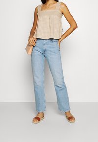 Top senza maniche color beige chiaro con scollatura quadrata e spalline arricciate, abbinato a jeans dritti blu chiaro e sandali piatti marroni.