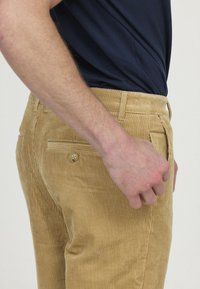 Pantalon en velours côtelé beige avec des côtes verticales, poche arrière boutonnée, taille ajustée et poches latérales, vu de côté.