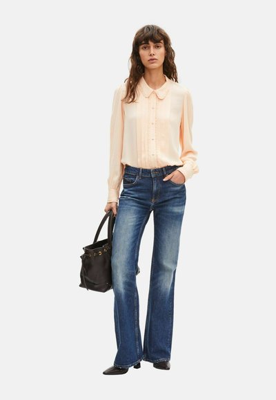 Mujer con blusa de manga larga color durazno pálido, jeans acampanados azules, zapatos negros puntiagudos, sosteniendo un bolso negro con una mano y con la otra en el bolsillo.