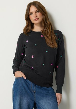 Femme aux longs cheveux bruns portant un pull noir avec des motifs d'étoiles roses et vertes et un jean bleu, debout avec une main dans la poche.