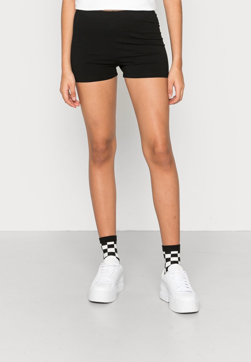 Personne portant un short noir ajusté, des chaussettes à carreaux noir et blanc, et des baskets blanches à plateforme, debout sur un sol gris clair.