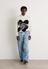 Suéter blanco con diseño floral negro, usado sobre una camisa con cuello, combinado con jeans de pierna ancha de color azul claro y zapatos negros. Conjunto simple y texturizado.