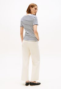 Sieviete stāv sāniski, valkājot baltu un tumši zilu svītru īsroku kreklu, krēmkrāsas platajiem biksēm ar frayed malām un melniem dzīvokļu apaviem.