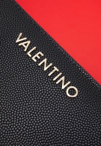 Crni kožni novčanik s teksturom koji prikazuje urezani zlatni logotip "VALENTINO." Pozadina je u solidnoj crvenoj boji.