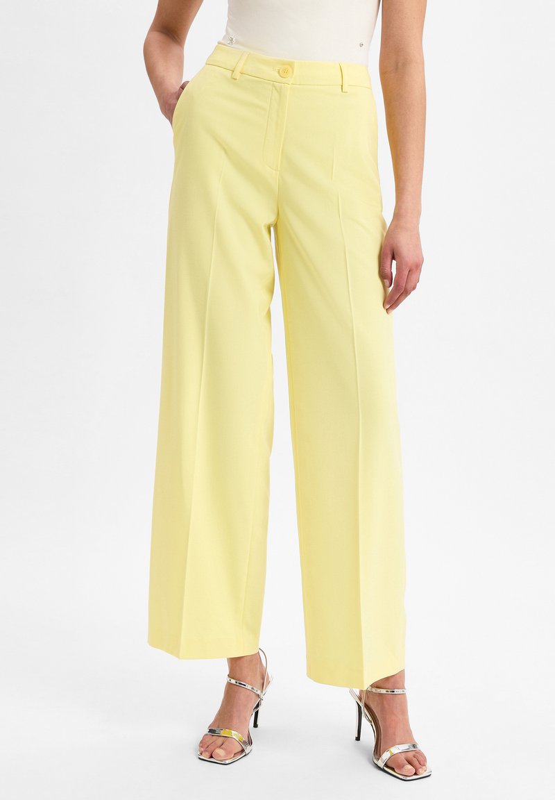 Heldere gele wijde broek met een gladde textuur, hoge taille, zijzakken en een enkele knoopsluiting aan de voorkant. Gecombineerd met metallic hakken.