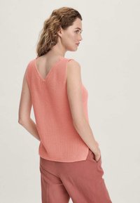 Pfirsichfarbener Stricktanktop mit V-förmigem Rücken, strukturiertem Gewebe und lockerer Passform. Kombiniert mit lockeren, hellrosa Hosen.