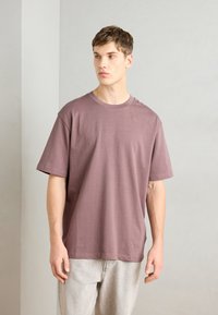 T-shirt de manga curta, com decote redondo em malva, feita de tecido suave. Apresenta um corte descontraído e uma textura suave, combinada com jeans de cor clara.