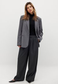 Grijze op maat gemaakte blazer met scherpe revers, gecombineerd met hoge grijze broek en een aansluitende zwarte coltrui. Strak, minimalistisch ontwerp.