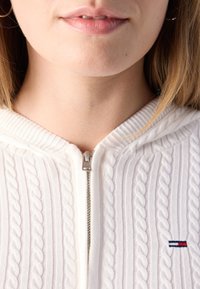 Maglione bianco lavorato a maglia con motivo a treccia, chiusura zip davanti e piccolo logo ricamato con accenti rossi, bianchi e blu. Tessuto texturizzato.