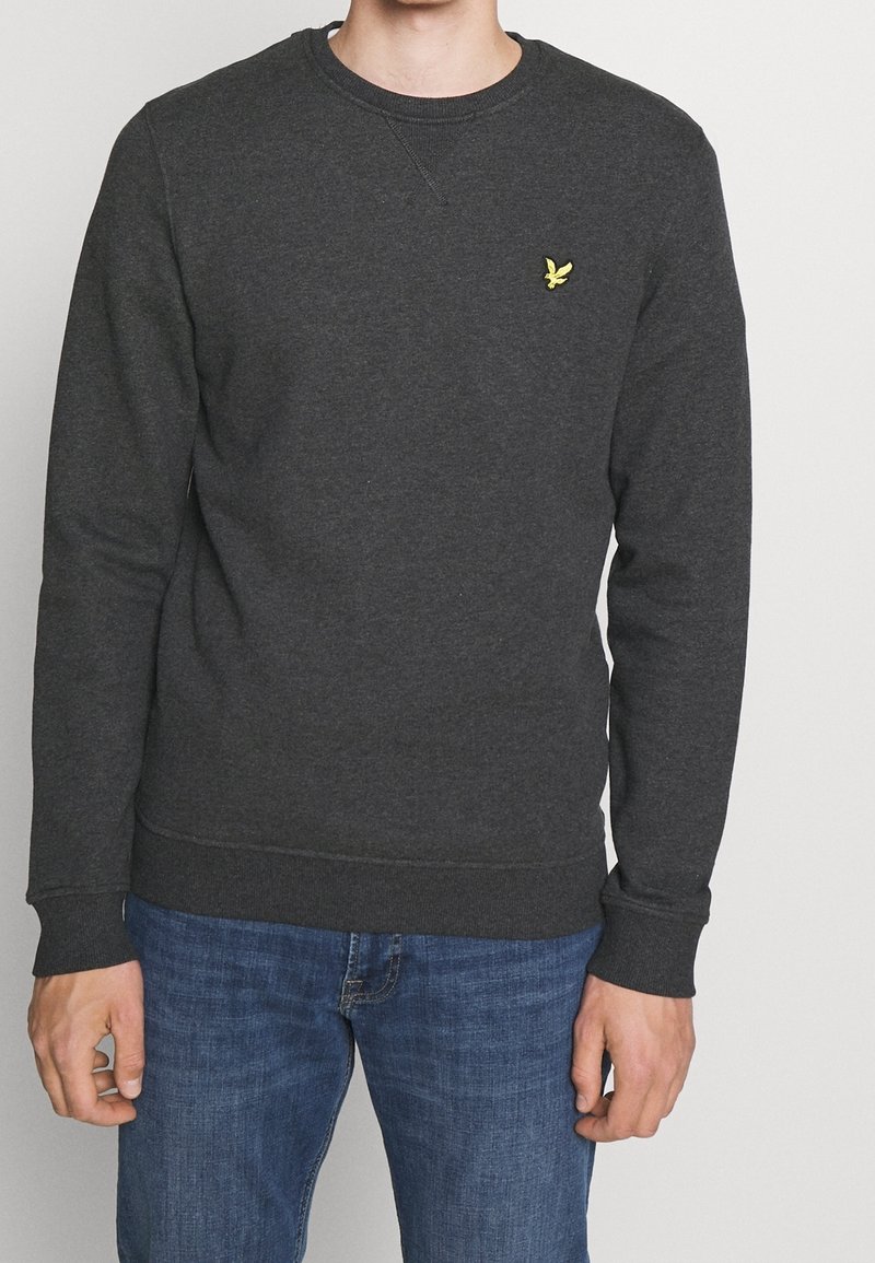Homme portant un sweat-shirt gris foncé à manches longues avec un petit logo jaune d'oiseau sur la poitrine et un jean bleu, debout devant un fond blanc.
