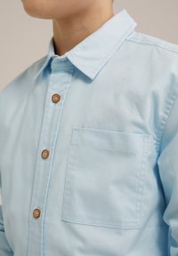 Světle modrá košile na knoflíky vyrobená z bavlněné tkaniny. Má klasický límec, hnědé knoflíky a náprsní kapsu. Hladká textura.