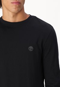 Mustkunst väikese halli puu logoga vasakul rinnal. Sile tekstuur, tavaline istuvus.
