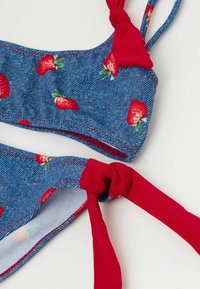 Top de bikini con patrón de mezclilla azul y lazos rojos, con pequeños estampados de fresas. Textura suave con bordes cosidos.