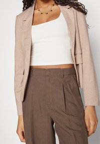 Veste beige cintrée avec un bouton unique, en tissu léger. Portée sur un crop top blanc et associée à un pantalon marron taille haute.