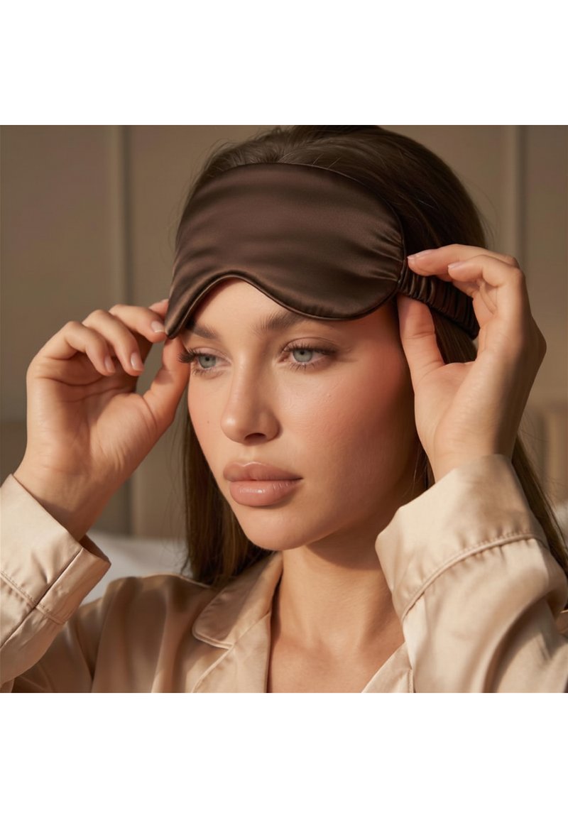 Giovane donna con lunghi capelli castani che indossa un pigiama beige aggiusta una maschera da sonno marrone scuro sulla fronte all'interno.