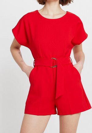 Femme portant une combinaison-short rouge à manches courtes avec ceinture et poches, debout les mains dans les poches devant un fond uni.