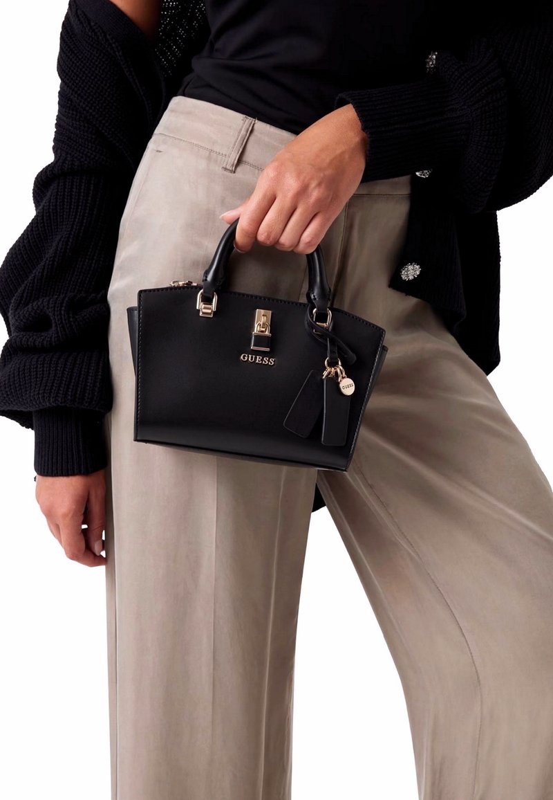 Borsa a tracolla sintetica nera con forma strutturata, hardware dorato e logo. Presenta doppi manici superiori e un accento a forma di portachiavi. Indossata con pantaloni chiari.