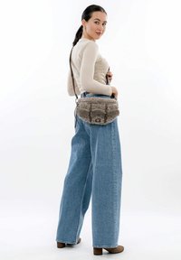 Donna in top beige con texture, jeans a gamba larga blu e stivali marroni, con una borsa a tracolla grigia e pelosa, in posa su sfondo bianco.
