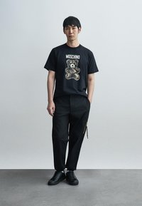 Μαύρο t-shirt Moschino με γραφικό αρκουδάκι, συνδυασμένο με χαλαρά μαύρα παντελόνια και μαύρα αθλητικά παπούτσια. Λεπτό ύφασμα, μινιμαλιστικός σχεδιασμός.