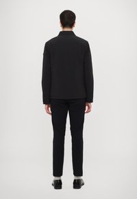 Jachetă neagră cu guler, închidere cu fermoar și mâneci lungi, asortată cu pantaloni închiși la culoare și pantofi, văzută din spate. Design monocrom.