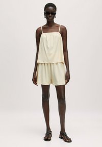 Lichtcrème tanktop met dunne bandjes en gelaagde uitstraling, gecombineerd met bijpassende shorts. De stof heeft een gestructureerde afwerking; het model draagt zwarte zonnebril.