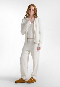 Set da loungewear in maglia testurizzata color crema, con cappuccio con zip, top aderente e pantaloni larghi. Pantofole marroni con dettaglio della cintura e fibbia.