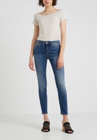 Světle béžový top s krátkým rukávem bez rukávů v kombinaci s tmavě modrými skinny džíny. Džíny mají vybledlý vzhled a černé a béžové otevřené boty na podpatku s třásněmi.