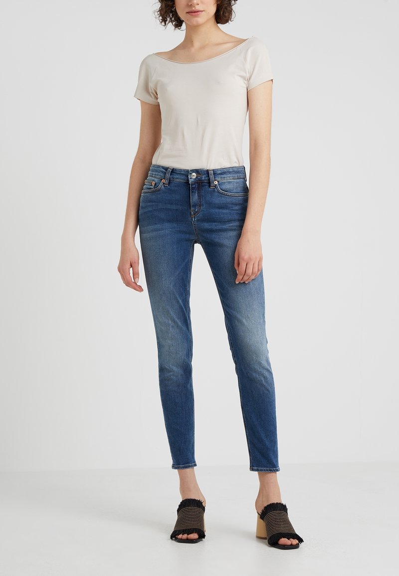 Světle béžový top s krátkým rukávem bez rukávů v kombinaci s tmavě modrými skinny džíny. Džíny mají vybledlý vzhled a černé a béžové otevřené boty na podpatku s třásněmi.