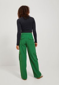 Groene cargobroek met zijzakken en metalen knopen, gecombineerd met een aansluitend donker topje. De broek heeft een wijd uitlopende pasvorm en een gladde textuur.