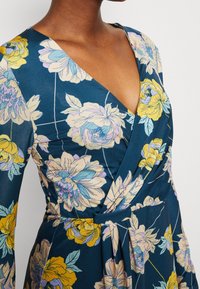 Robe portefeuille florale en marine avec des fleurs jaunes et bleu clair. Elle présente un décolleté en V profond, des manches longues et une taille froncée avec un détail à nouer.