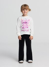 Biała bluza z długim rękawem z różowym nadrukiem kreskówkowej postaci, zestawiona z czarnymi dzwonami z sztruksu i szarymi butami wsuwanymi.