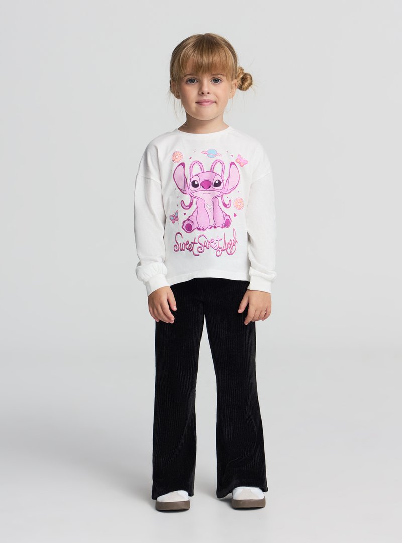 Biała bluza z długim rękawem z różowym nadrukiem kreskówkowej postaci, zestawiona z czarnymi dzwonami z sztruksu i szarymi butami wsuwanymi.