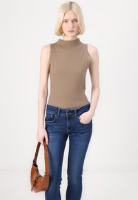Rjava brezrokavnik s visokim ovratnikom, v kombinaciji z modrimi jeans hlačami. Model drži majhno rjavo torbico z teksturirano površino.