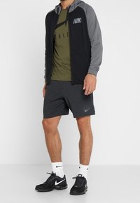 Manlig träningsoutfit bestående av en svart och grå huva zipjacka, en olivgrön t-shirt, grå shorts och svarta Nike-sneakers.