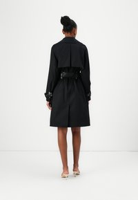 Zwarte trenchcoat met een aansluitende pasvorm, grote kraag en een ceintuur in de taille. Voorzien van een oversized rugflap en met knopen sluitende manchetten. Gladde textuur van de stof.