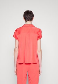Korallfärgad blus med korta ärmar och krage, med en baktill för att ge extra rörelse och flöde. Slät tyg med en mjuk glans.