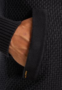 Maglione nero con texture, caratterizzato da polsini a coste e una tasca laterale. La tasca presenta un piccolo etichetta arancione che dettaglia il marchio.