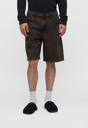 Man draagt donkerbruine knielange shorts, witte kousen, zwarte instapschoenen en een donker lange mouwen top, staand tegen een effen achtergrond.