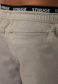Beige broek met een gladde textuur, voorzien van een zwarte tailleband met "STHUGE" in witte letters. Bevat een klein logo-detail op de achterkant.