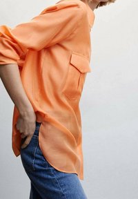 Maglietta a maniche lunghe arancione con taschino sul petto, in tessuto leggero e con una vestibilità ariosa, abbinata a jeans in denim blu e una silhouette rilassata.
