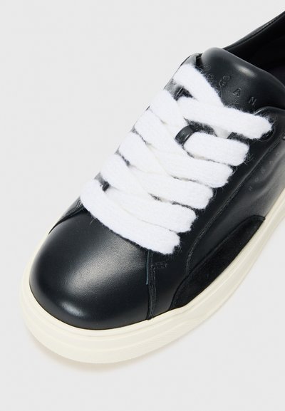 Black leather sneaker with white textured laces, cream rubber sole, rounded toe, and subtle branding on the side. Smooth and polished finish.
