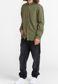 Camisa de botones de manga larga de color verde oliva, con rayas verticales, un único bolsillo en el pecho y botones marrones, combinada con jeans negros y zapatillas blancas.