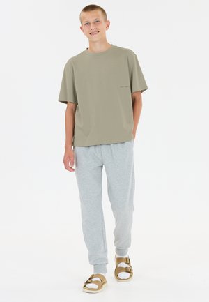 Teenagedreng smilende, iført olivengrøn oversized t-shirt, lysegrå joggingbukser, hvide sokker og lysebrune sandaler, stående mod en hvid baggrund.