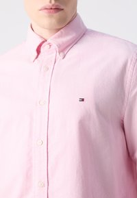 Gaiši rozā pogājamā krekla, kas izgatavots no kokvilnas. Tam ir klasiskas formas apkakle, baltas pogas un neliels izšūts logo uz krūšu.