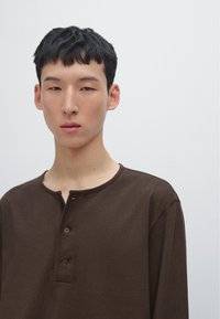 Braunes Langarmhemd mit rundem Ausschnitt, versehen mit einer Knopfleiste und minimalen Nähdetails. Glatte Stofftextur.