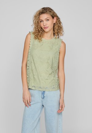 Svetlo zelena čipkasta majica s designom zakrivljenih rukava, zapletenimi diamantnimi vzorci in sproščenim krojem, v kombinaciji s svetlo modrimi jeans hlačami.
