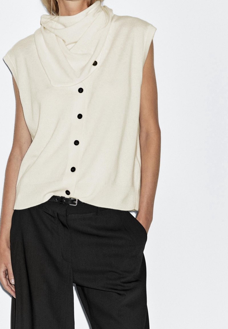 Femme portant un haut crème sans manches boutonné avec col plié et un pantalon noir à larges jambes avec ceinture, debout, main dans la poche.