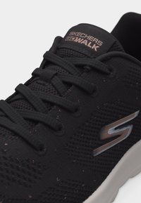 Čierna teniska Skechers GOwalk s textilnou sieťovinou, čiernymi šnúrkami a bielou podrážkou, na boku so znakom "S" od Skechers.