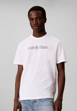 Młody mężczyzna ubrany w białą koszulkę Calvin Klein i jasnoniebieskie dżinsy, stojący na jednolitym szarym tle.